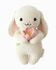 The Cuddle + Kind Baby Bunny (Blooming Tulip) is a cream-colored, heirloom-quality Peruvian cotton plush with floppy ears, black eyes, a sweet smile, and holds a pink flower with a yellow center in its arms.