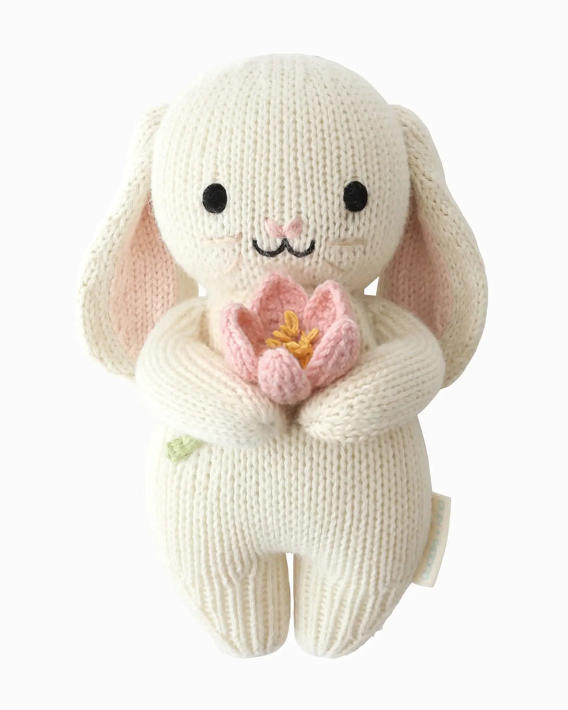 The Cuddle + Kind Baby Bunny (Blooming Tulip) is a cream-colored, heirloom-quality Peruvian cotton plush with floppy ears, black eyes, a sweet smile, and holds a pink flower with a yellow center in its arms.