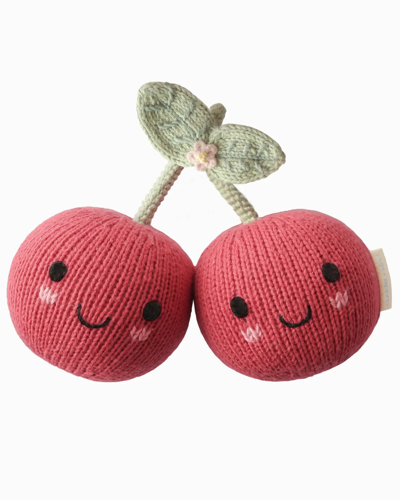 The Cuddle + Kind Baby Cherry features two pink knitted cherries with smiling faces, blushing cheeks, and green hand-knit stems with leaves, all beautifully handcrafted in a plush style from heirloom-quality natural Peruvian cotton yarn.