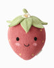 The Cuddle + Kind Baby Strawberry is a knitted plush strawberry, crafted from soft Peruvian cotton yarn, featuring a green top, a small white flower, and a smiling face with rosy cheeks on a plain white background—an heirloom-quality treasure inspired by hand-knit baby animals.