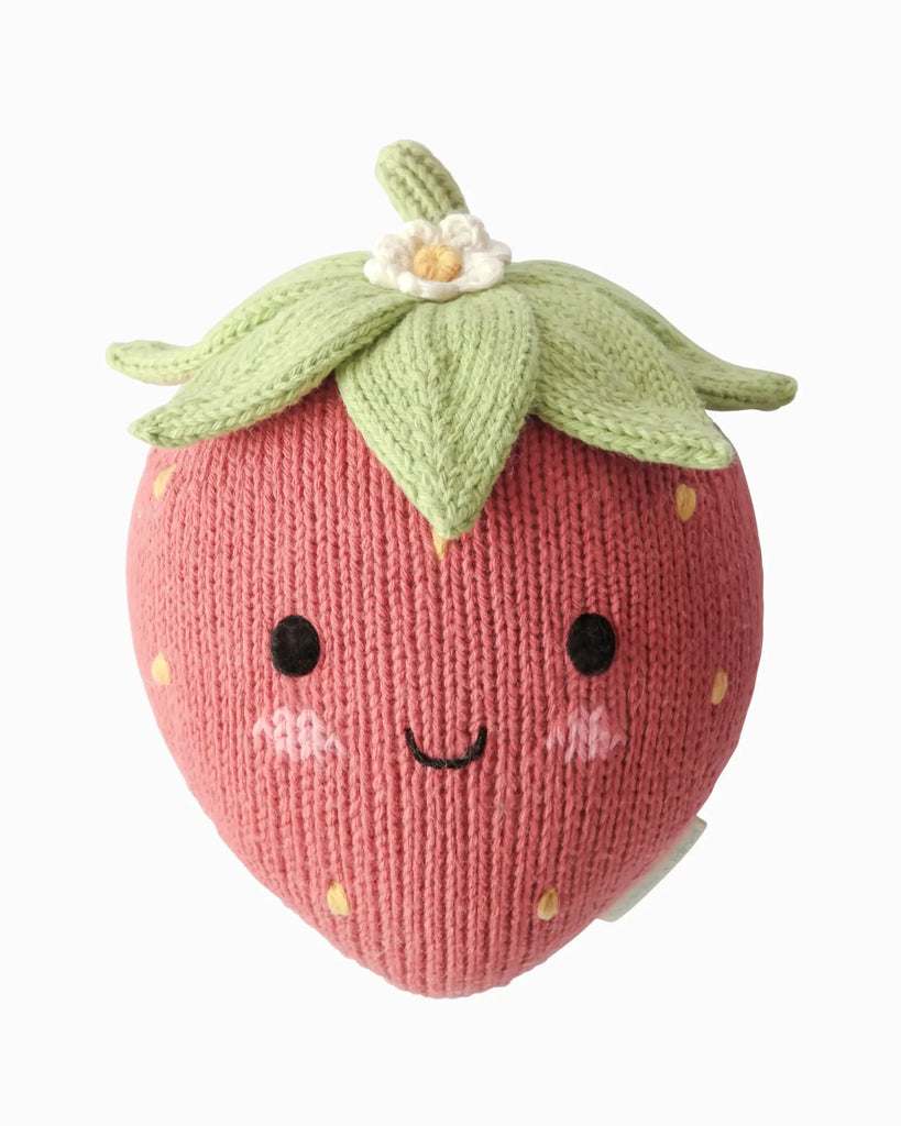 The Cuddle + Kind Baby Strawberry is a knitted plush strawberry, crafted from soft Peruvian cotton yarn, featuring a green top, a small white flower, and a smiling face with rosy cheeks on a plain white background—an heirloom-quality treasure inspired by hand-knit baby animals.