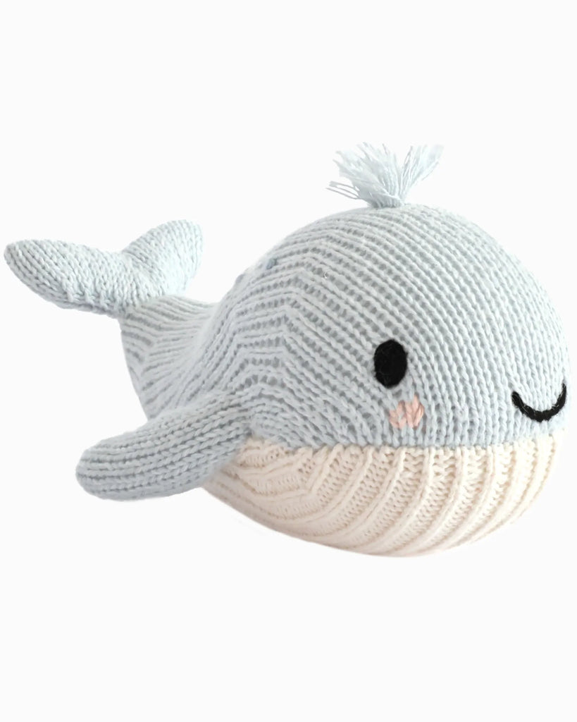 The Cuddle + Kind Baby Whale is a soft, knitted plush toy shaped like a light blue whale with a smiling face, black eyes, pink blush, and a textured white belly—handmade in Peru.