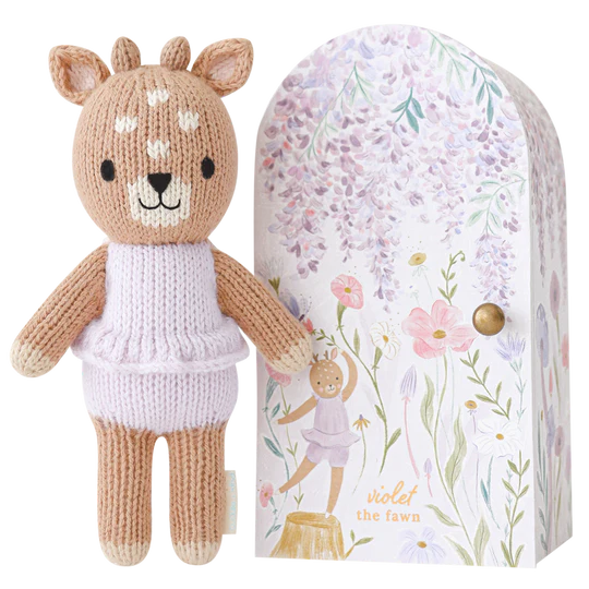 Cuddle + Kind Tiny Violet The Fawn toy standing next to a floral illustrated packaging with the name "Tiny Violet the fawn." The toy is beige and white, wearing a knitted dress, and the box features delicate