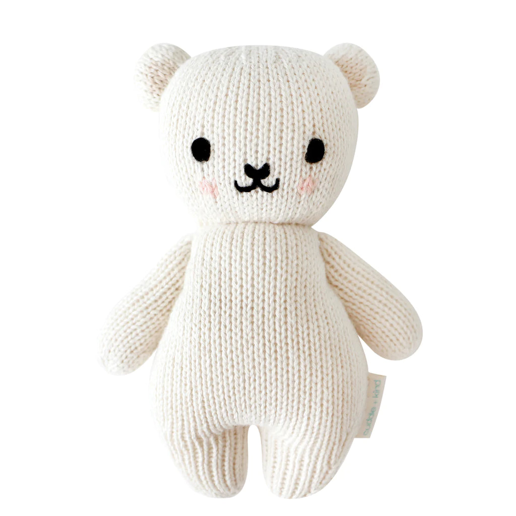 A Cuddle + Kind Baby Polar Bear with a cream-colored body, black eyes, a small black stitched nose, and a simple black mouth. Its cheeks are marked with light pink blush. Hand-knit from natural Peruvian cotton yarn, this heirloom-quality bear has small, rounded ears and appears soft and cuddly.