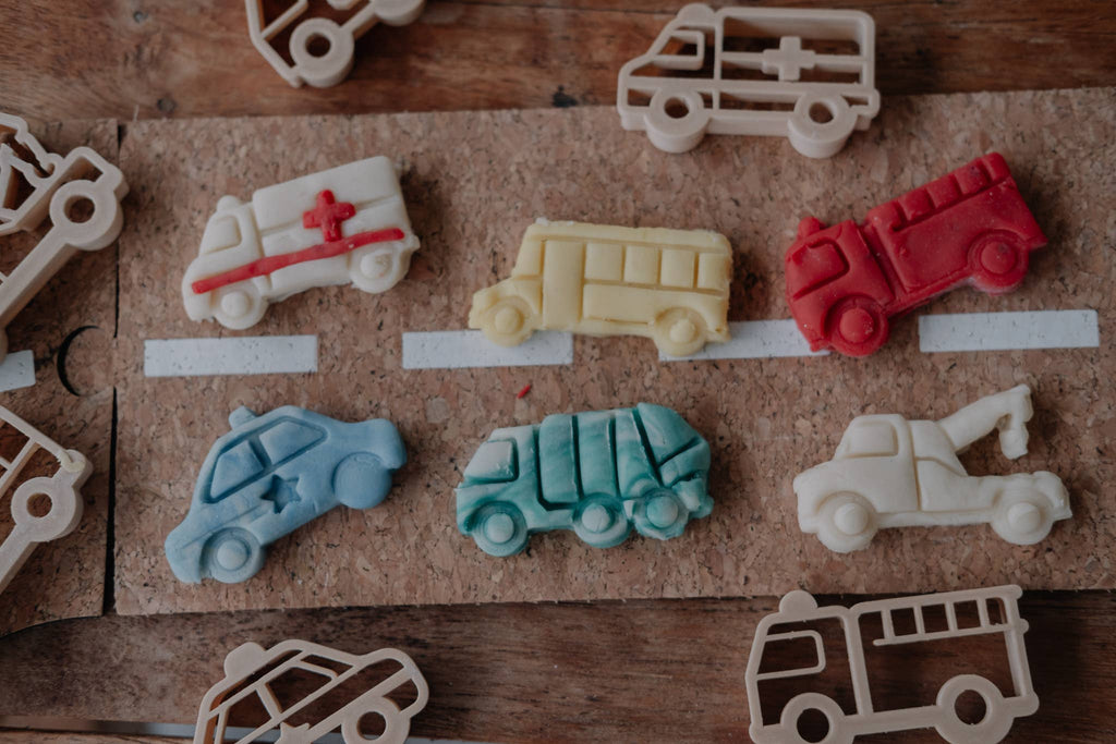 Clay toy vehicles—including a fire truck, school bus, ambulance, and cars—are lined up on a cork board road with hand-drawn lanes. The Service Vehicle Cutter Set - Eco Playdough Tool is displayed nearby, inspiring imaginative sensory play with its transport-themed design.