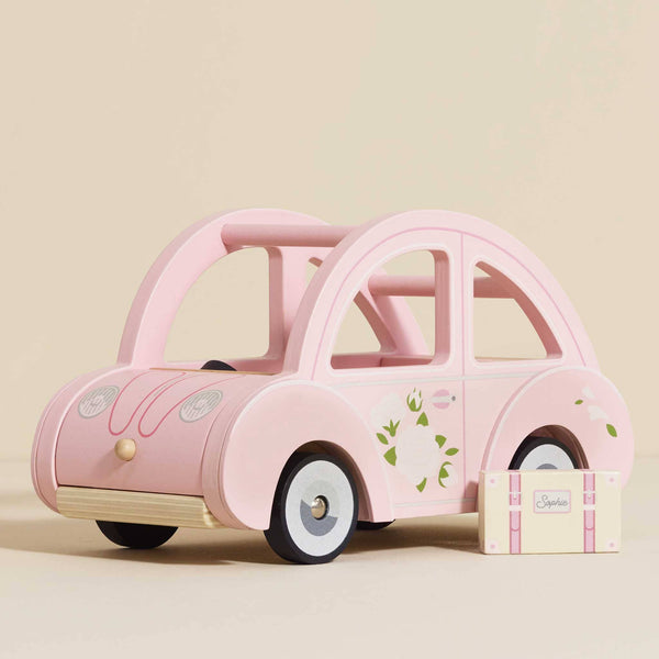 Sophie's Dolls House Toy Car—a pink retro wooden car with floral designs—sits beside a matching "Sophie" suitcase on a beige background, ideal as an educational toy or charming nursery decor.