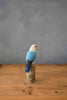 The Hand Carved Ornament - Lovebird - Light Blue - 7.5"H features a realistic blue and white lovebird perched on sustainable softwood, displayed on a wooden surface with a simple gray background.