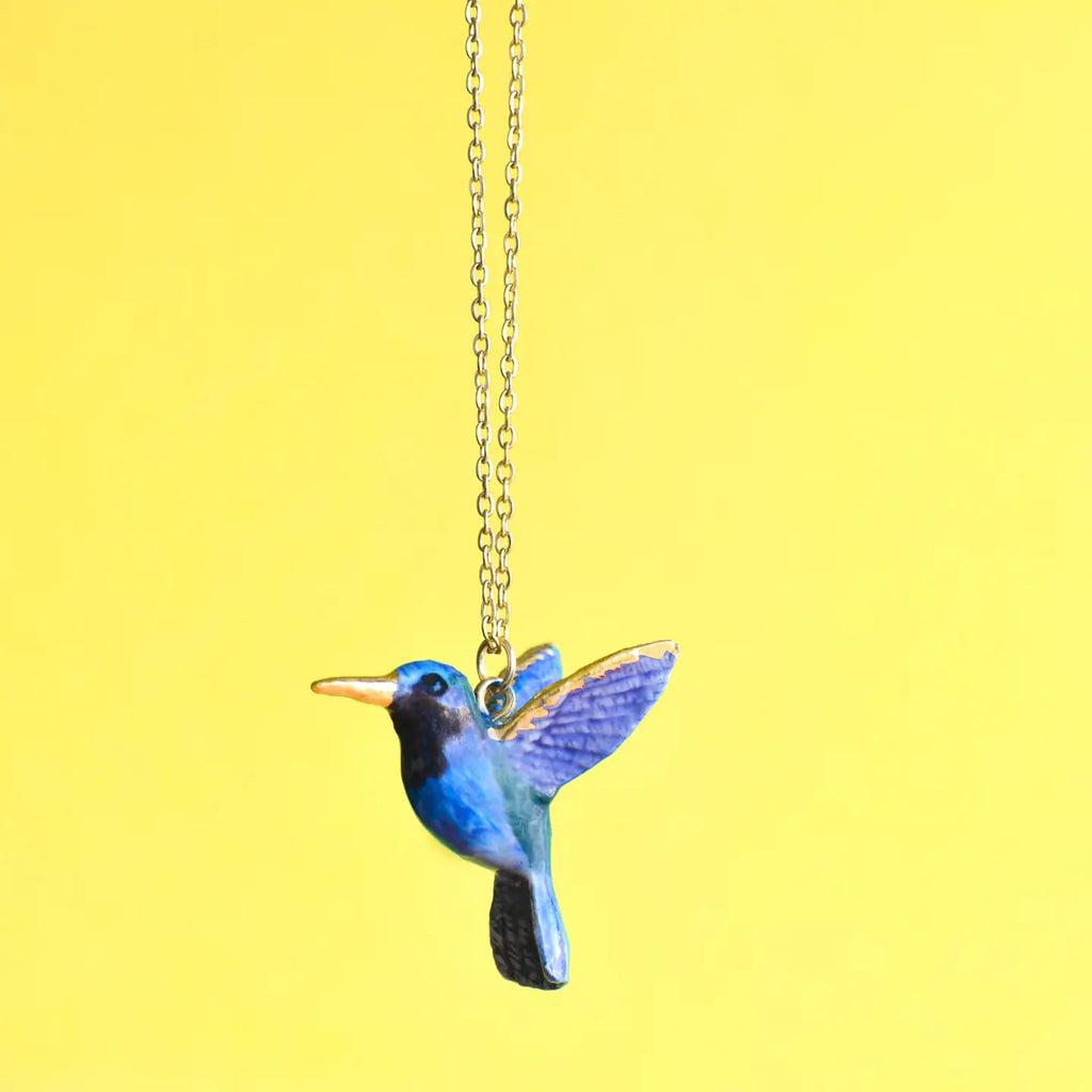 A Hummingbird Necklace features a detailed pendant with blue and black plumage, hand-painted feathers, and gold accents. The 24k gold plated hummingbird appears to be in flight against a solid, vibrant yellow background.