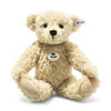 A 12-inch Steiff Luca Teddy Bear, light brown and plush with black eyes, a black nose, and the signature Steiff "Button in Ear" tag in its left ear, sits upright with arms open and legs slightly spread.
