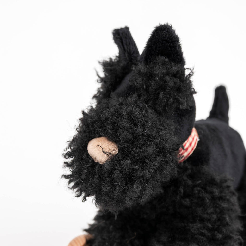 A close-up of Pull-Along Walter, a black stuffed dog with curly fur, a beige nose, and a red-striped collar, sits atop wooden wheels like an adorable pull-along toy against a white background.