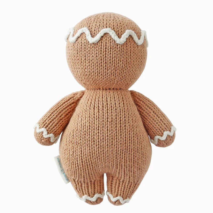 The Cuddle + Kind Baby Gingerbread toy, made from heirloom-quality natural Peruvian cotton yarn, is adorned with white icing-like trimming on its head, arms, and legs. This charming piece is set against a white background and evokes the timeless appeal of hand-knit baby animals treasured for generations.