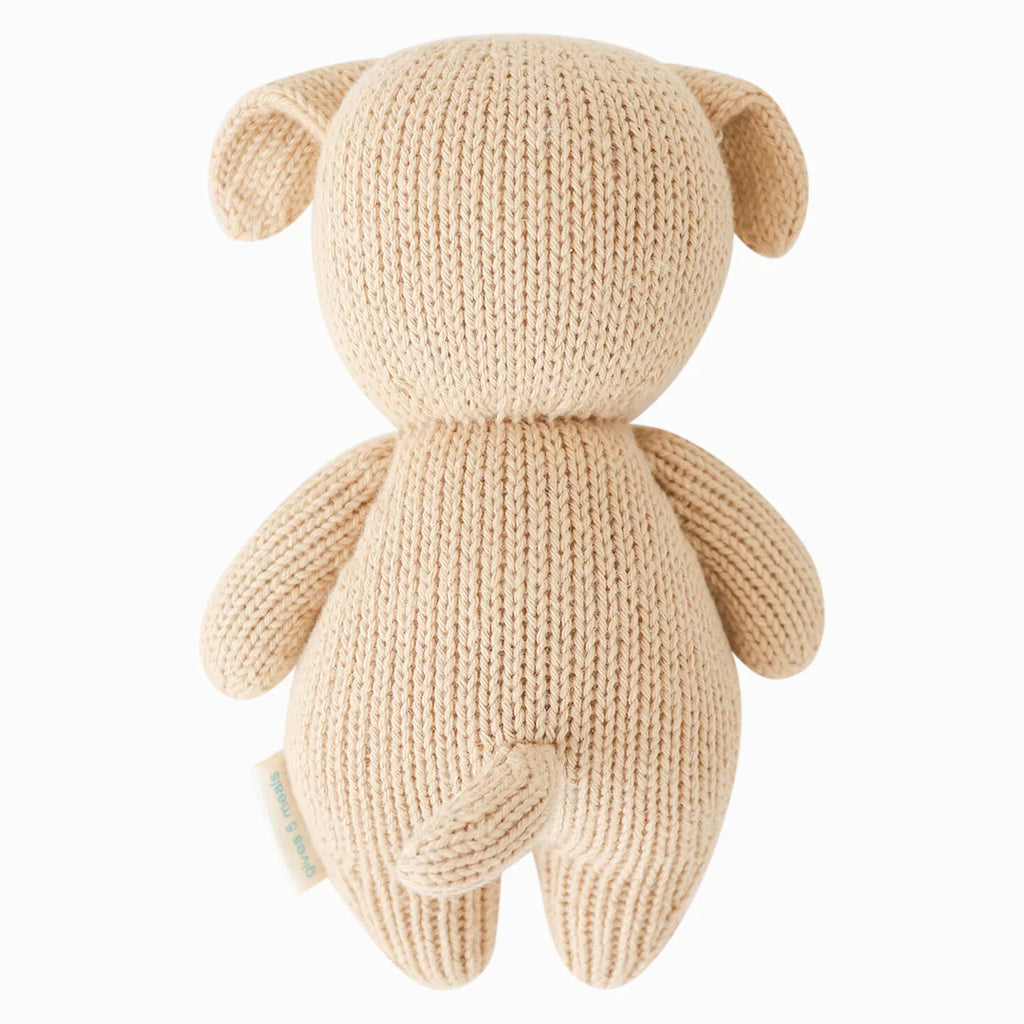 A hand-knit beige Cuddle + Kind Baby Puppy facing away with visible seams, featuring rounded ears and limbs, crafted from Peruvian cotton yarn. It is posed against a white background and has a small fabric tag.