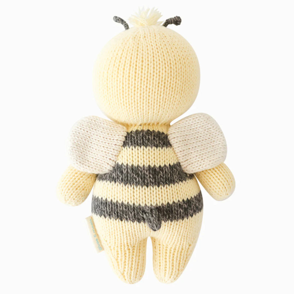 A hand-knit Cuddle + Kind Baby Bee resembling a bee with yellow and black stripes on its body, white wings, and details, crafted from Peruvian cotton yarn and displayed against a white background.