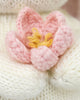 A close-up of the Cuddle + Kind Baby Bunny (Blooming Tulip) features a soft, knitted pink flower with yellow center details, crafted from heirloom-quality Peruvian cotton yarn, resting on a white knitted surface.