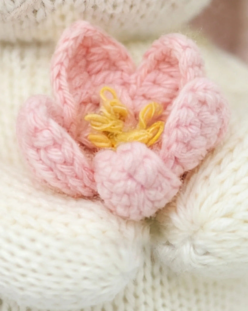 A close-up of the Cuddle + Kind Baby Bunny (Blooming Tulip) features a soft, knitted pink flower with yellow center details, crafted from heirloom-quality Peruvian cotton yarn, resting on a white knitted surface.