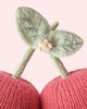 Close-up of the Cuddle + Kind Baby Cherry, featuring two pink knitted cherries with green stems and leaves, handcrafted from natural Peruvian cotton yarn and adorned with a small pink knitted flower, displayed against a pale pink background.