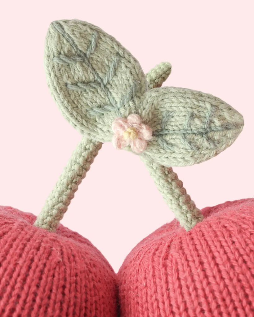 Close-up of the Cuddle + Kind Baby Cherry, featuring two pink knitted cherries with green stems and leaves, handcrafted from natural Peruvian cotton yarn and adorned with a small pink knitted flower, displayed against a pale pink background.
