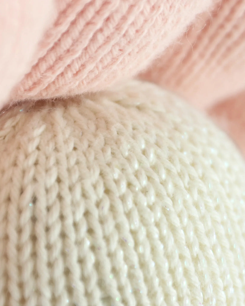 Close-up of Cuddle + Kind Baby Pearl shows soft, heirloom-quality knitted fabric in pale pink and off-white, highlighting the texture and pattern of natural cotton yarn.