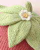 Close-up of the Cuddle + Kind Baby Strawberry knitted item, featuring green leaves, a small white and yellow flower, and a green stem on a pink, textured background. Lovingly crafted from heirloom-quality Peruvian cotton yarn, this piece showcases exquisite attention to detail and softness.