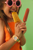 A smiling child in pink flower sunglasses and an orange top holds two popsicles, showing off her colorful Mini Kyomo Watch - Watercolor against a green background.