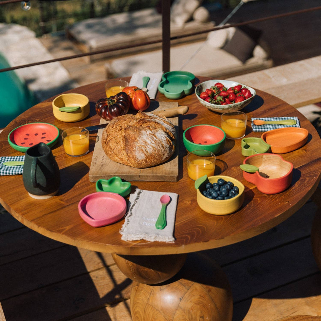 A round wooden table is set outdoors with a loaf of bread, fruit bowls, juice, colorful plates—including the Cathy the Carrot Plate & Spoon Set—cups, and utensils. The spread features strawberries, blueberries, tomatoes, napkins arranged for brunch, and non-toxic baby tableware specially designed for little guests.