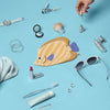 A hand reaches for a dropper bottle among beauty products, jewelry, sunglasses, a toothbrush, and the Butterfly Fish Pencil Case, all arranged on a light blue background.
