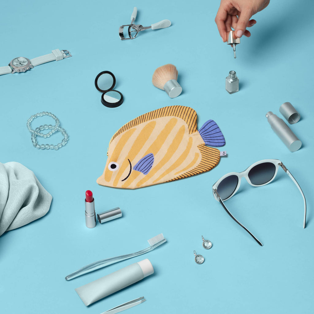 A hand reaches for a dropper bottle among beauty products, jewelry, sunglasses, a toothbrush, and the Butterfly Fish Pencil Case, all arranged on a light blue background.