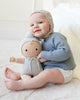 A baby in a blue sweater sits on a soft carpet, smiling and hugging the Cuddle + Kind My First Baby (Bluebell) doll, dressed in light-colored clothes; ships in one week.