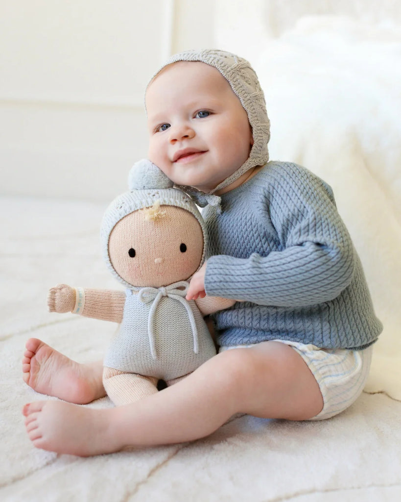 A baby in a blue sweater sits on a soft carpet, smiling and hugging the Cuddle + Kind My First Baby (Bluebell) doll, dressed in light-colored clothes; ships in one week.