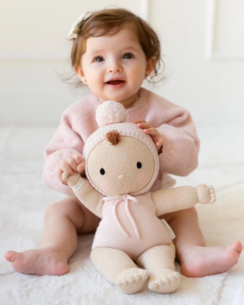 A smiling toddler with light brown hair in a pink sweater and white bow sits on a soft surface, holding the Cuddle + Kind My First Baby (Blush)—a hand-knit doll in a matching pink outfit with pom-pom hat, made from natural cotton yarn. Ships in one week.