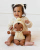 A smiling baby with pigtails and a cream bow sits wearing a cream sweater, holding the Cuddle + Kind My First Baby (Buttercup), a hand-knit, fair trade baby doll made from natural cotton yarn, featuring a pom-pom hat and outstretched arms. Ships in one week.