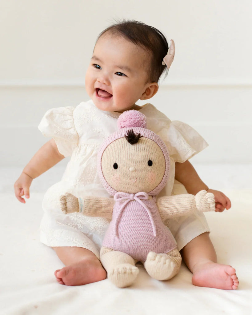 A smiling baby with a bow hair clip sits on a light surface, holding the Cuddle + Kind My First Baby (Lavender) doll—hand-knit from natural cotton yarn with a pink outfit and hat. Ships in one week. The scene is happy and playful.