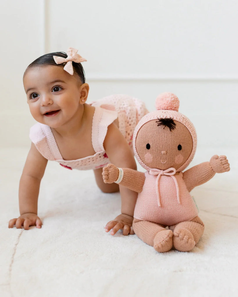 A smiling baby in a pink dress crawls on the floor next to the Cuddle + Kind My First Baby (Peach), a hand-knit doll made from natural cotton yarn with a pink pom-pom hat—ships in one week.