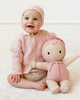 A smiling baby in a pink knit bonnet and sweater sits holding the Cuddle + Kind My First Baby (Rose), a hand-knit doll in a pom-pom hat and bow. Both wear light, natural cotton outfits. Doll ships in one week.