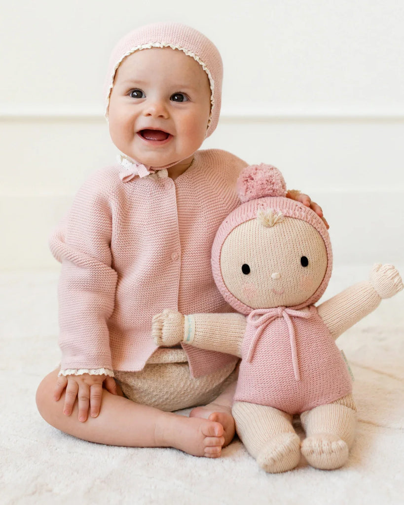 A smiling baby in a pink knit bonnet and sweater sits holding the Cuddle + Kind My First Baby (Rose), a hand-knit doll in a pom-pom hat and bow. Both wear light, natural cotton outfits. Doll ships in one week.