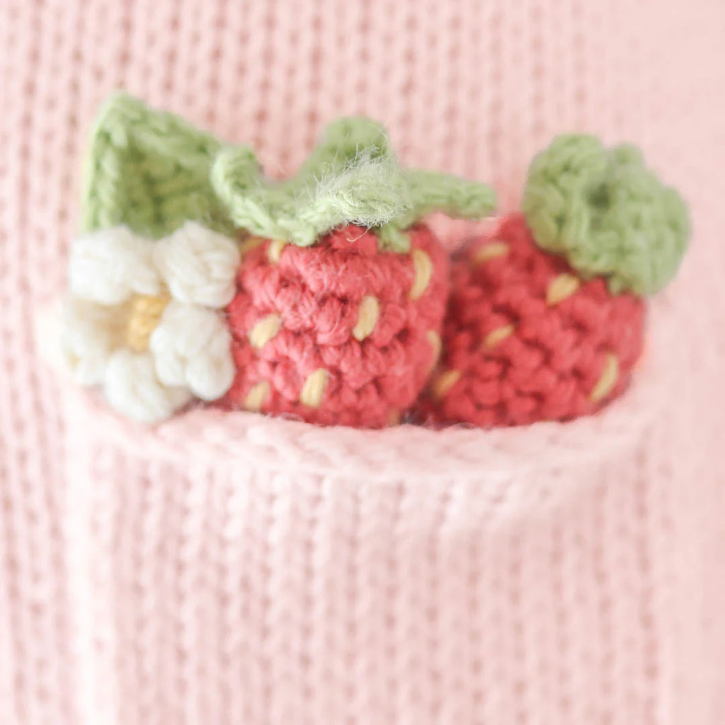 Close-up of Cuddle + Kind Ava the Cow - Powder Pink, handmade in Peru from premium cotton yarn, with a small pocket holding two crocheted strawberries and a white flower with yellow center.
