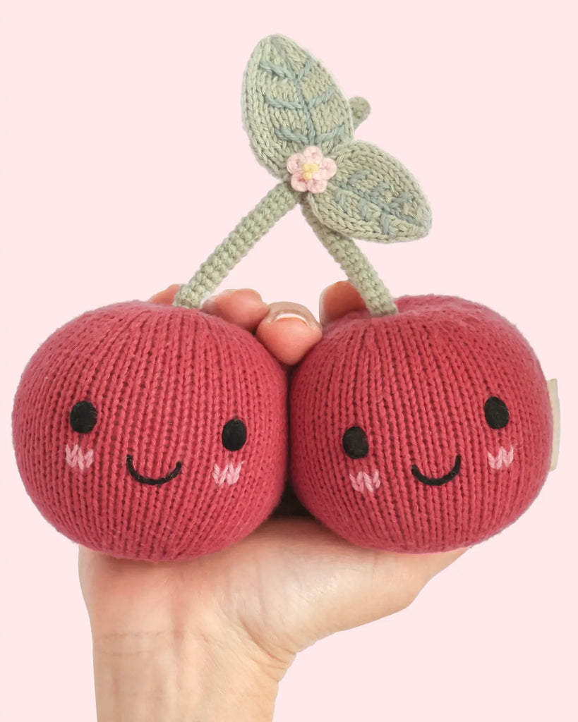A hand holds two Cuddle + Kind Baby Cherry plushies, each featuring smiling faces, blushing cheeks, green leaves, and a tiny pink flower on top. Handmade from natural Peruvian cotton yarn for an heirloom-quality finish, these sweet designs stand out against a light pink background.