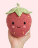A hand holds the Cuddle + Kind Baby Strawberry, an heirloom-quality knitted plush toy crafted from soft Peruvian cotton yarn, featuring a cute smiling face, blushing cheeks, and green leaves on top, against a light pink background.