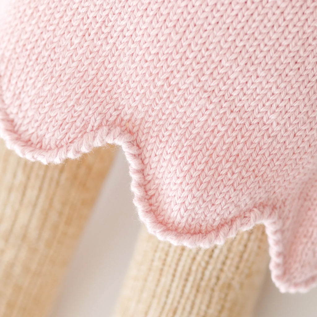 Close-up of Cuddle + Kind Bella the Honey Bear’s pink sustainable fair trade knitted fabric with a scalloped edge, draped over two light beige ribbed cylinders. The textures of both fabrics are clearly visible.