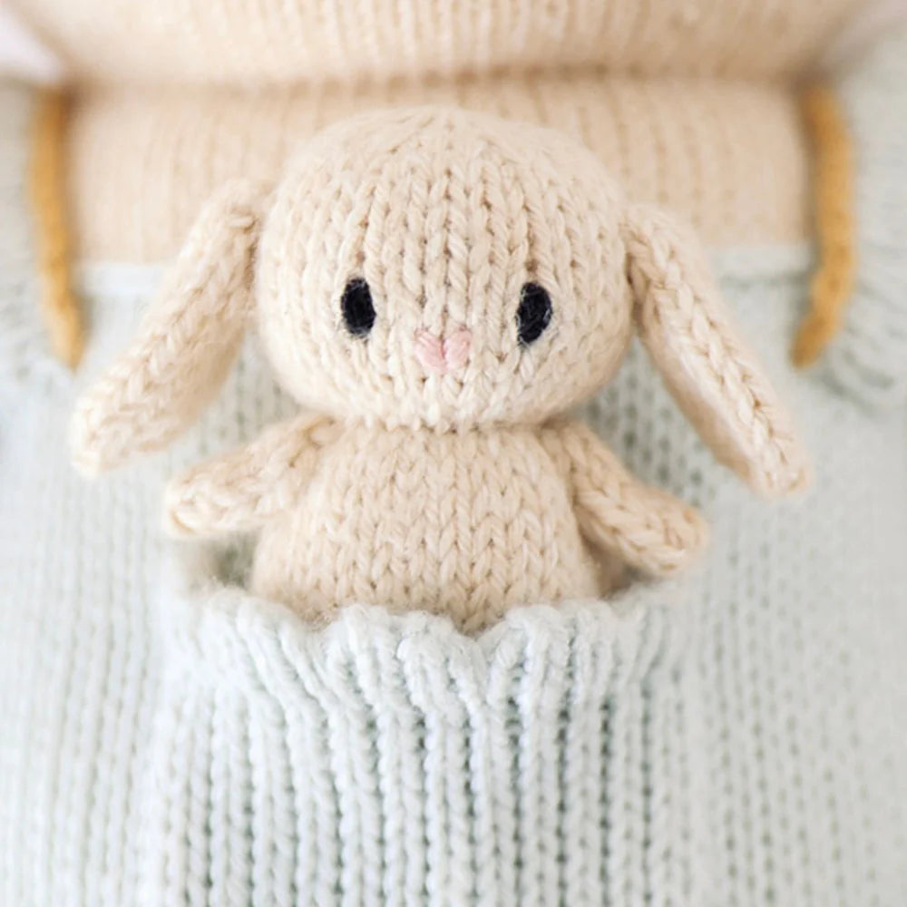 Cuddle + Kind Briar the Bunny – Light Blue is a small, cream-colored hand-knit doll with long ears, nestled in the front pocket of a light blue cotton yarn garment.