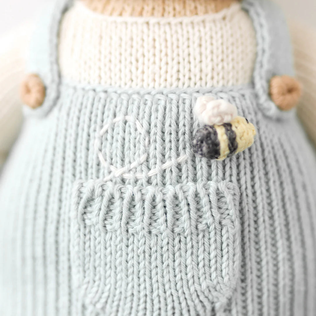 Close-up of a Cuddle + Kind the Honey Bear doll wearing a light blue dress, featuring delicate textures and a small white flower with a yellow center attached to the front.