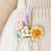 A close-up of a Cuddle + Kind the Honey Bear adorned with small, colorful crochet flowers and leaves, featuring soft pastel shades of yellow, pink, blue, and green.