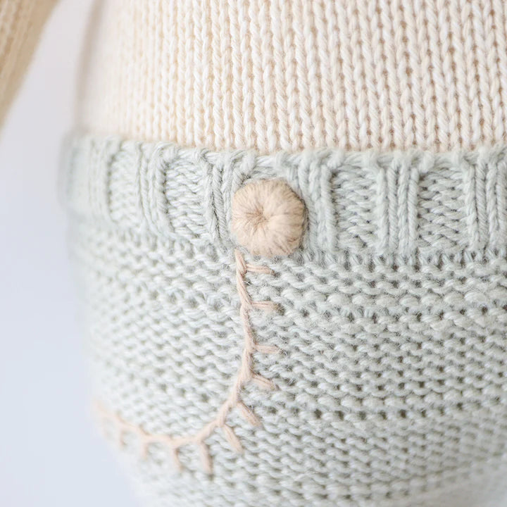 Close-up of a Cuddle + Kind Sawyer The Lion - Large featuring varying textures and colors, including shades of beige and green. A prominent beige knitted button and a loose thread detail are visible, crafted from Peruvian cotton yarn.