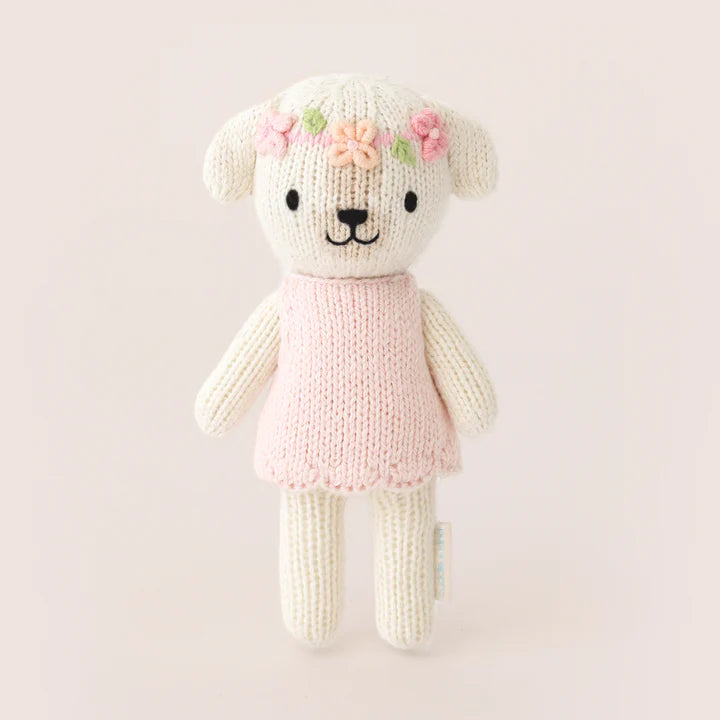 A handmade knitted toy lamb wearing a pink dress, adorned with floral accents on its head, standing against a plain light background, crafted following fair trade practices: Cuddle + Kind Tiny Charlotte The Dog.