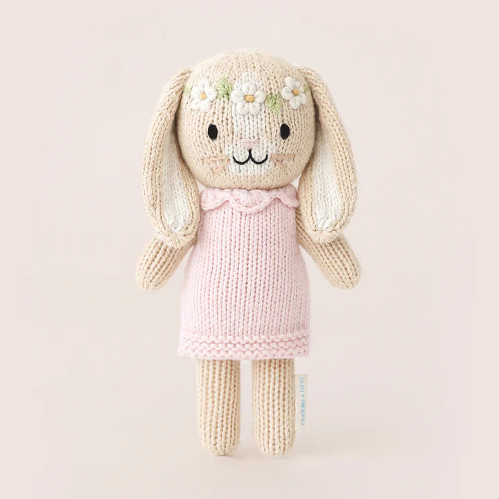 A Cuddle + Kind Tiny Hannah The Bunny hand-knit, cream-colored bunny doll wearing a light pink dress. The non-toxic bunny has embroidered facial features and a garland of white flowers stitched on its head. It stands upright against a plain, light background, embodying fair trade practices.