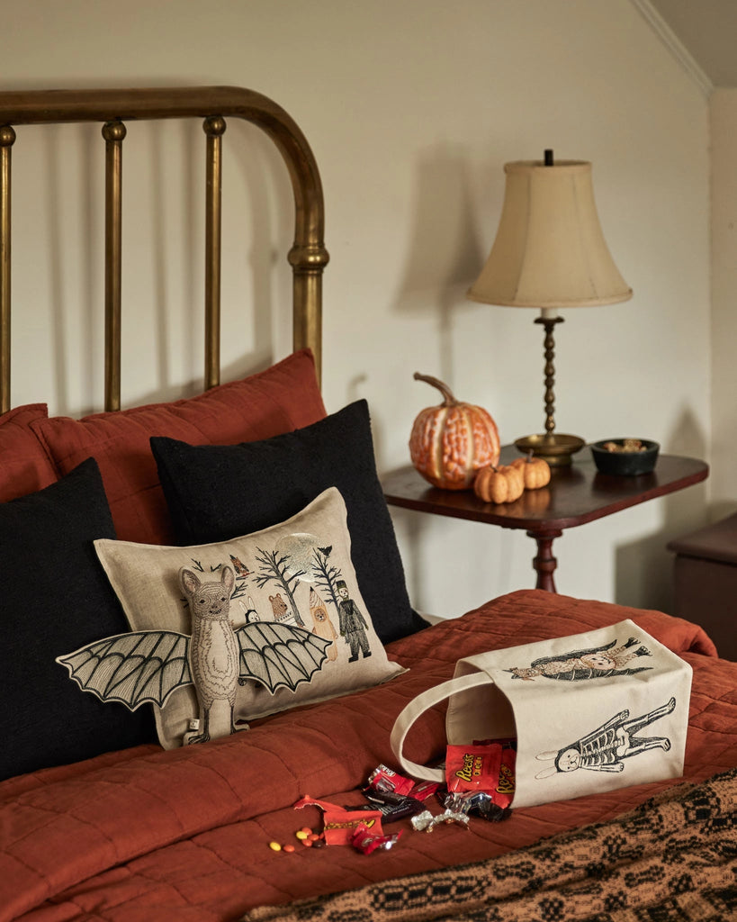 A cozy bed with rust-colored bedding and Halloween pillows, including a bat design. On the bed is a Coral & Tusk Halloween Trick Or Treat Bag (ships in one week) filled with candy; pumpkins and a lamp sit on the bedside table.