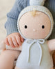 A child's hand gently holds the Cuddle + Kind My First Baby (Bluebell), a soft, hand-knit doll in a pale blue hooded outfit with pom-pom, featuring a simple stitched face with black eyes and blushing cheeks. Ships in one week.