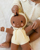 A baby in a cream knit outfit sits on a light surface, gently playing with the Cuddle + Kind My First Baby (Buttercup), a hand-knit cotton doll dressed in yellow with a tied bow under its chin. Ships in one week.