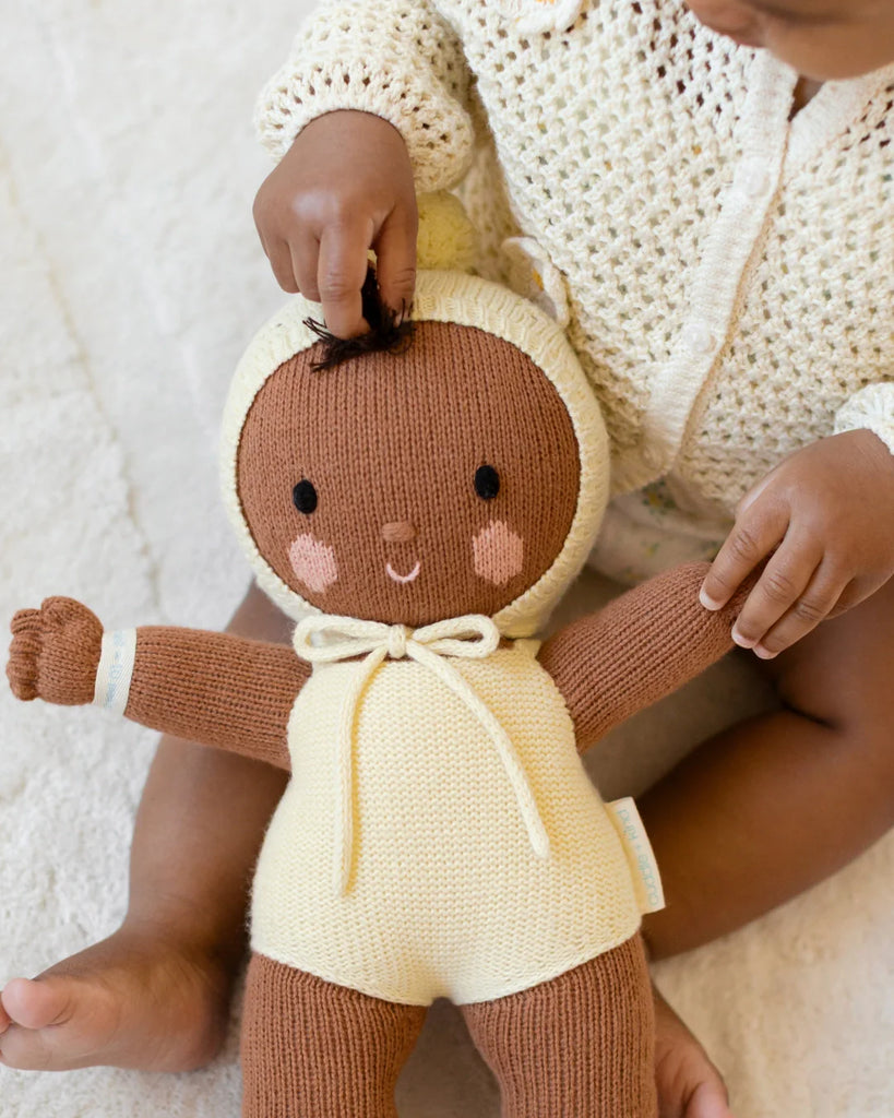 A baby in a cream knit outfit sits on a light surface, gently playing with the Cuddle + Kind My First Baby (Buttercup), a hand-knit cotton doll dressed in yellow with a tied bow under its chin. Ships in one week.