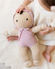 A child in a white dress sits on a soft surface, gently holding the Cuddle + Kind My First Baby (Lavender), a fair trade hand-knit doll dressed in a pink hooded outfit—ships in one week.